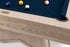 Close-up of a pool table corner showing a light natural wood frame, dark blue felt cloth, and several billiard balls including a yellow 8-ball, white cue ball, and orange ball.