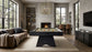 A modern living room featuring a dark wood pool table centered on a large beige rug, with a fireplace, large windows, a brown sofa, and a hanging brass chandelier.