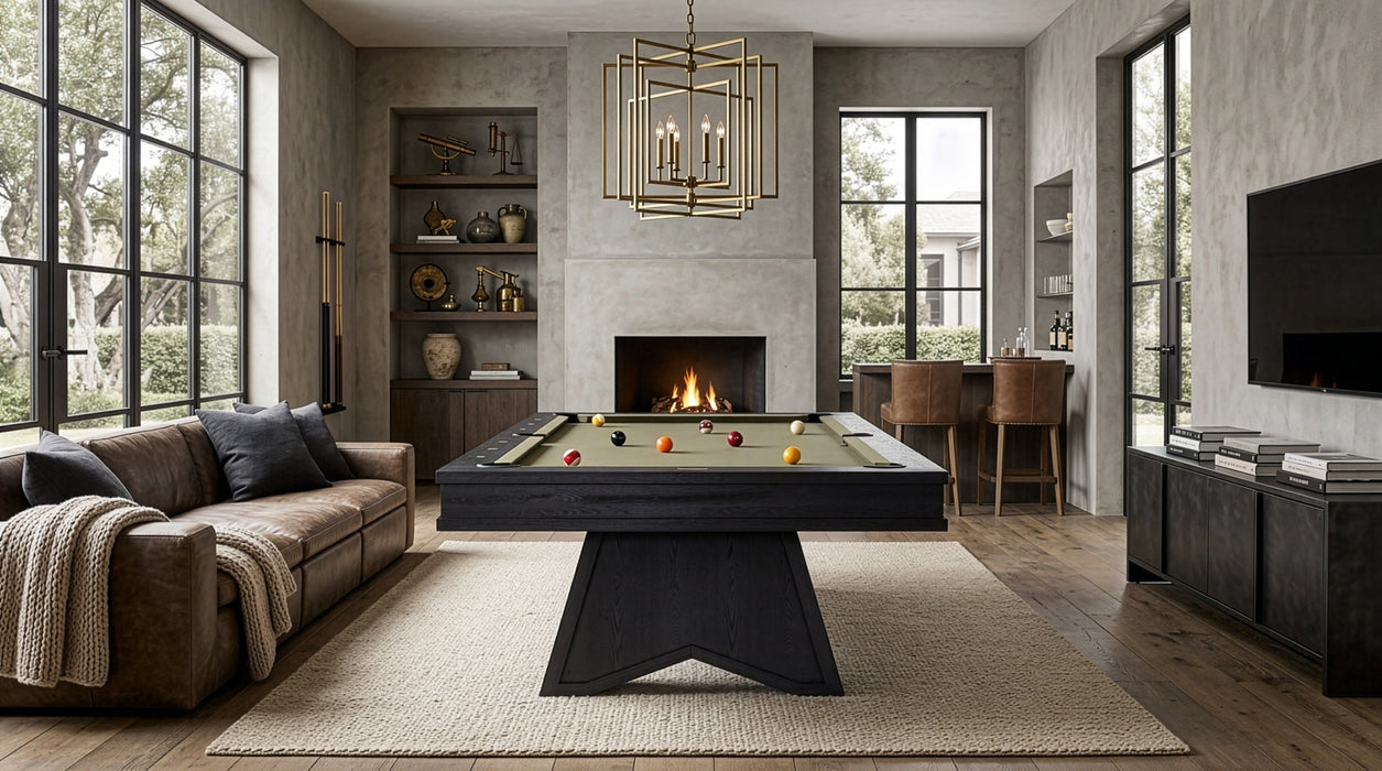A modern living room featuring a dark wood pool table centered on a large beige rug, with a fireplace, large windows, a brown sofa, and a hanging brass chandelier.