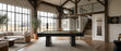 A modern dark wood pool table with a green felt surface is centered in a spacious, high-ceilinged rustic game room with exposed wood beams, large windows, a staircase, and a loft.