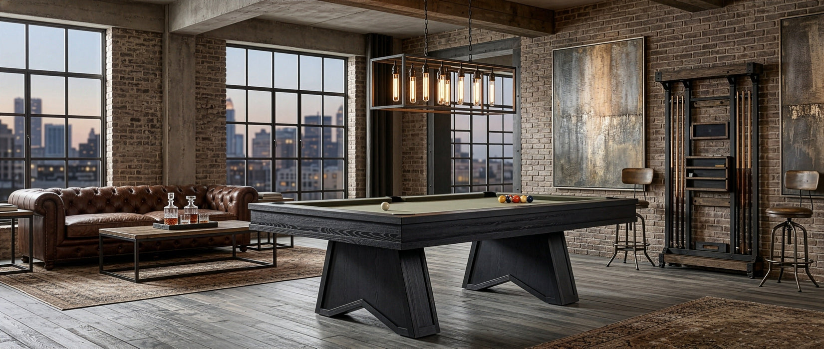 A dark wood pool table with a green felt surface is shown in a styled loft room with brick walls, large windows, a leather sofa, and a city skyline view.