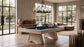 A light wood pool table with a deep blue felt surface is shown in a modern, sunlit living room with large windows, a sectional sofa, and a chandelier.