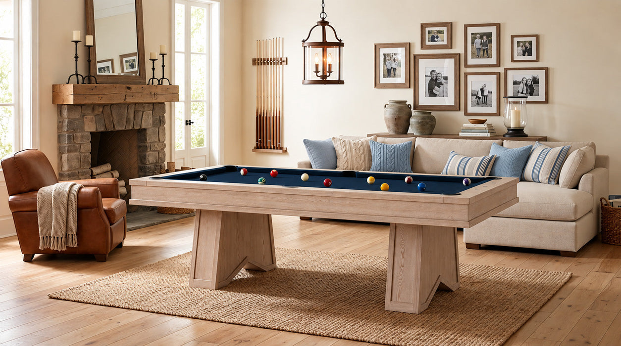 A light natural wood pool table with a blue felt playing surface and colorful billiard balls, set in a beautifully styled living room with a fireplace, sofa, and framed wall art.