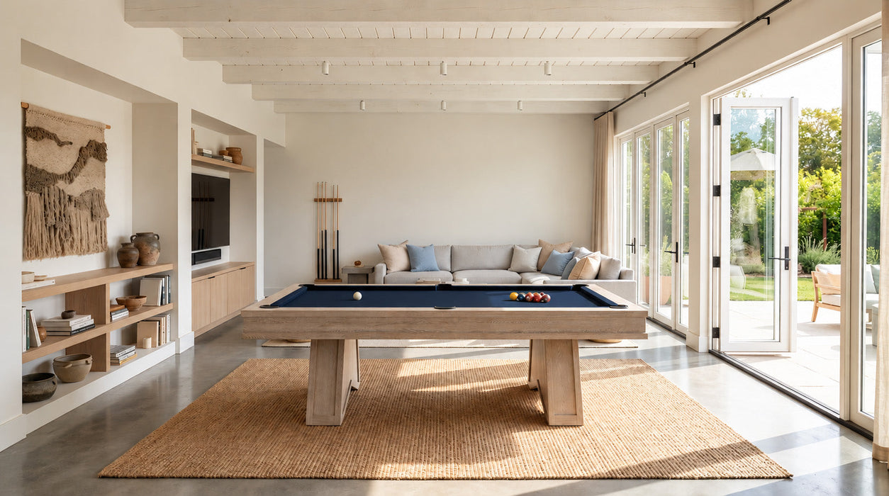 A modern wooden pool table with a blue felt surface is centered in a bright, contemporary room with large glass doors, a woven rug, and a light gray sofa in the background.