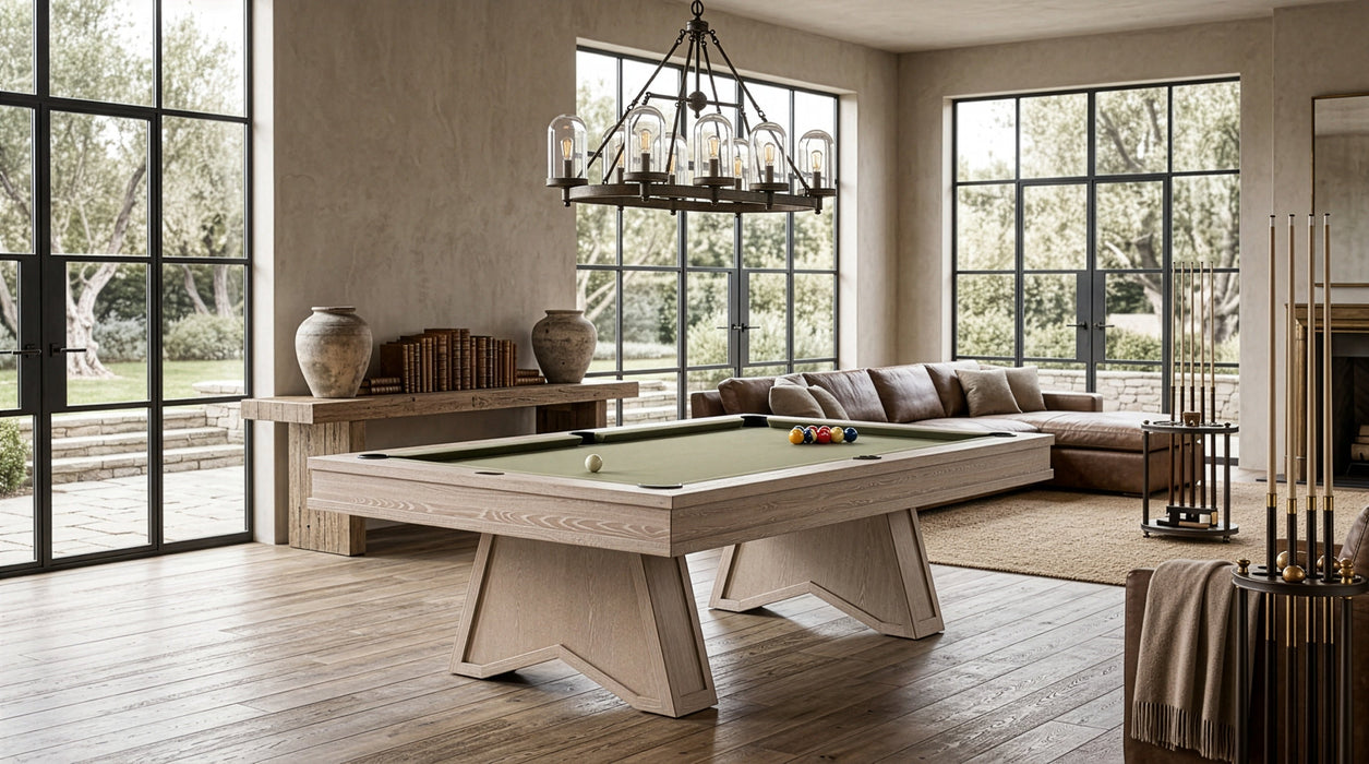 A light natural wood pool table with a green felt surface is shown in a modern, sunlit living room with large windows, a sectional sofa, and a chandelier.