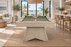 A light natural wood pool table with green felt sits in a bright, modern beach-view living room. The table is set on a large woven jute rug, with a rack of colorful billiard balls and a white cue ball on the green felt.