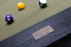 Close-up of a wooden billiards table corner with a brass plaque reading American Heritage Billiards, a green felt surface, and three pool balls nearby.