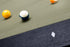 Close-up of a pool table with a green felt surface, dark wood edge, and billiard balls including a yellow ball, a blue-and-white ball, and part of an orange ball. A small white square object lies near the lower right edge.