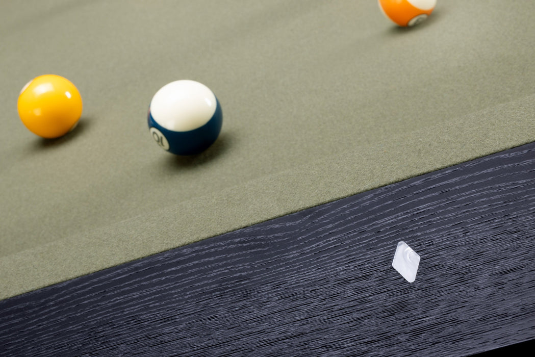 Close-up of a pool table with a green felt surface, dark wood edge, and billiard balls including a yellow ball, a blue-and-white ball, and part of an orange ball. A small white square object lies near the lower right edge.
